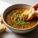 Quick lentil soup recipe with crusty bread, topped with fresh herbs in a white ceramic bowl on wooden surface.